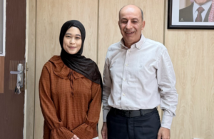 Two people smiling indoors against wood-paneled wall