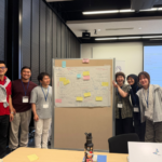 Six workshop participants pose beside a sticky-note brainstorming board.