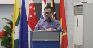 Man in batik shirt speaking at podium before flags