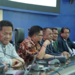 Officials seated at conference table with microphones and flags