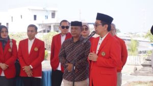 Pria berbicara di mikrofon bersama pejabat berjaket merah
