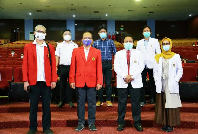 Masked professionals pose in auditorium wearing red blazers and white coats.
